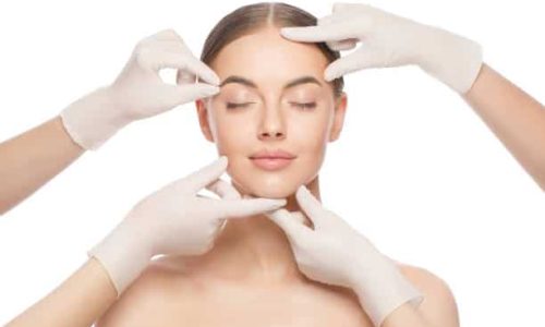 Close-up portrait of young woman standing with naked shoulders and neck, face is touched by beauticians in gloves, preparing her for plastic surgery procedures, isolated on white background